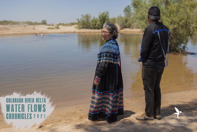 A woman in traditional Cucapá dress and young man stand near a body of water. The brown dirt is visible through the clear water and trees are in the background. Antonia Torres and son, Jaziel, visit the flowing Colorado River. June 2021.